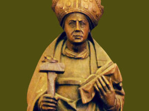 Wooden statue of a saint wearing a ornate crown and religious garments, holding a cross in one hand and a scroll in the other, set against a green background.